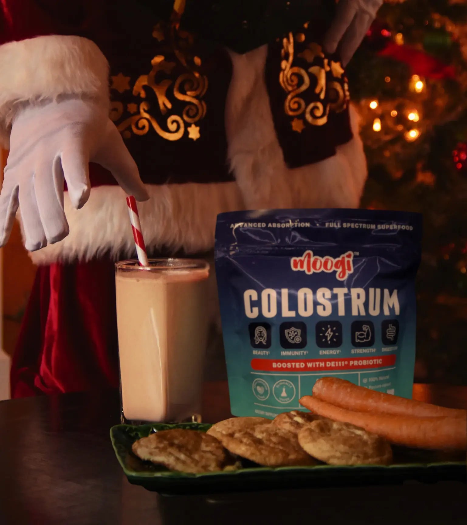 Festive holiday scene with Moogi colostrum supplement in blue packaging, a creamy drink, cookies, carrots, and Santa by a glowing Christmas tree, highlighting gut health, immunity, and beauty benefits.